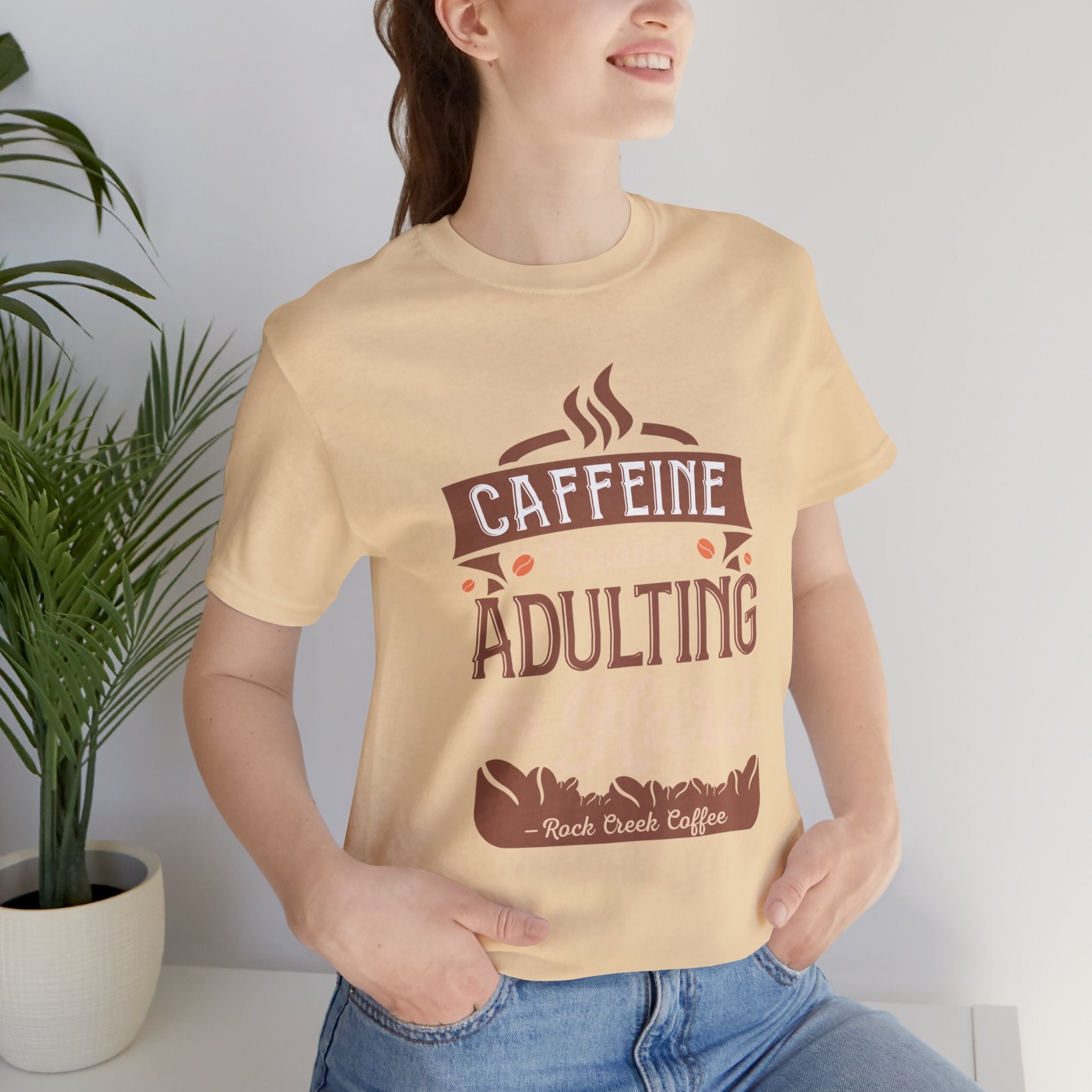 Adult woman modeling a soft cream short-sleeve T-shirt featuring the “Caffeine Because Adulting Is Hard” graphic in a stacked typographic design, photographed indoors beside a potted plant against a light neutral background.
