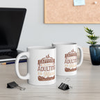 Two white Rock Creek Coffee mugs featuring the phrase “Caffeine Because Adulting Is Hard” in brown and tan lettering, displayed on a desk with a laptop, binder clips, books, and a small potted plant in the background.