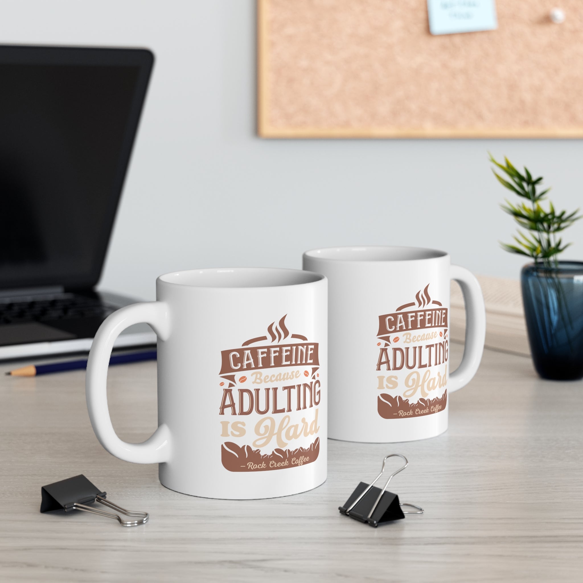 Two white Rock Creek Coffee mugs featuring the phrase “Caffeine Because Adulting Is Hard” in brown and tan lettering, displayed on a desk with a laptop, binder clips, books, and a small potted plant in the background.