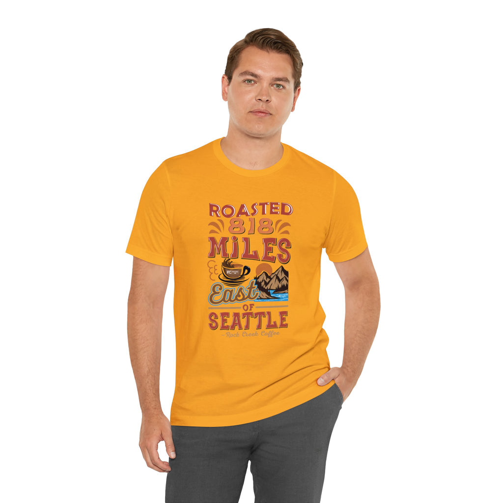 Adult man modeling a golden yellow short-sleeve T-shirt printed with a colorful graphic that reads “Roasted 818 Miles East of Seattle,” featuring a steaming coffee cup and mountain lake illustration, shown against a plain white background.