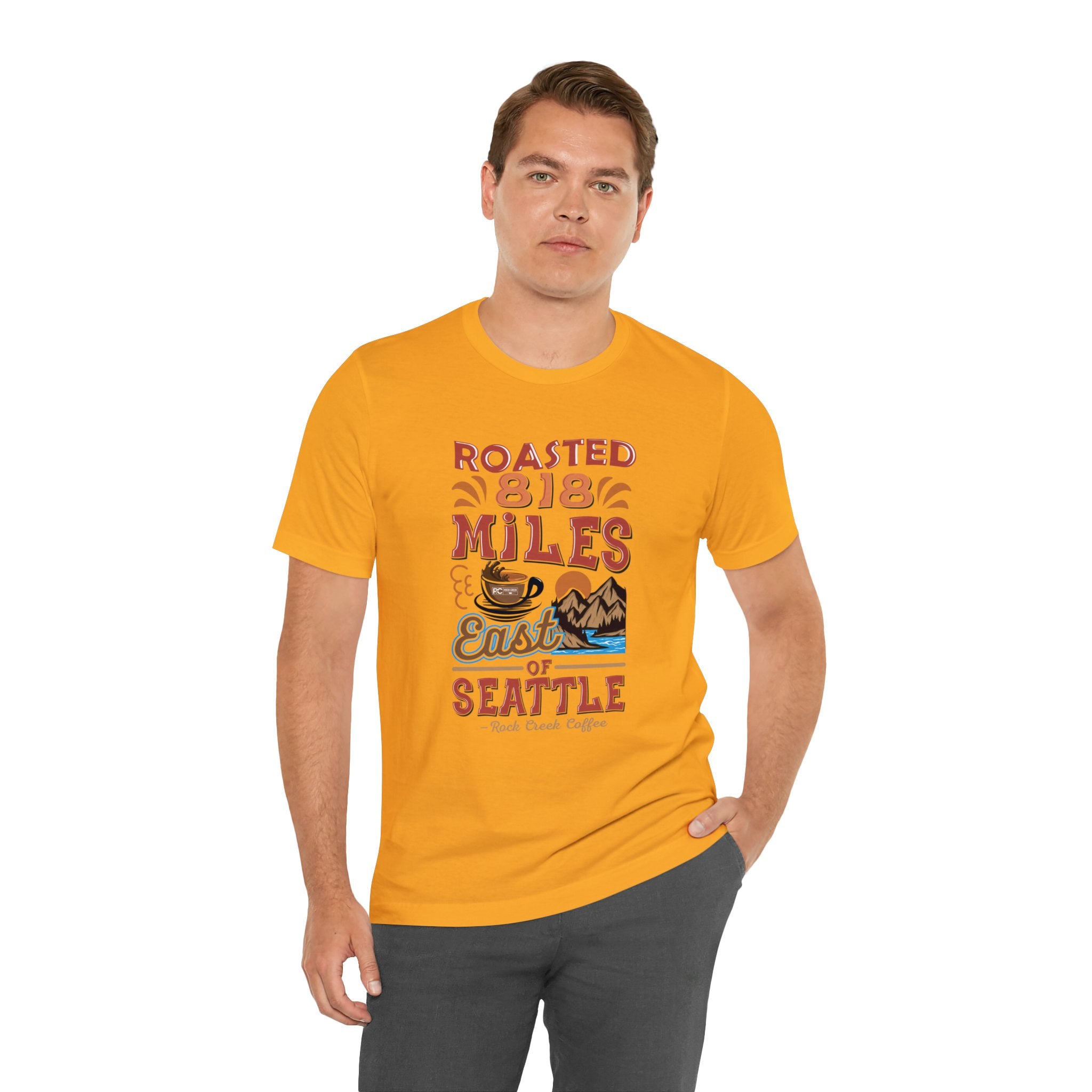 Adult man modeling a golden yellow short-sleeve T-shirt printed with a colorful graphic that reads “Roasted 818 Miles East of Seattle,” featuring a steaming coffee cup and mountain lake illustration, shown against a plain white background.