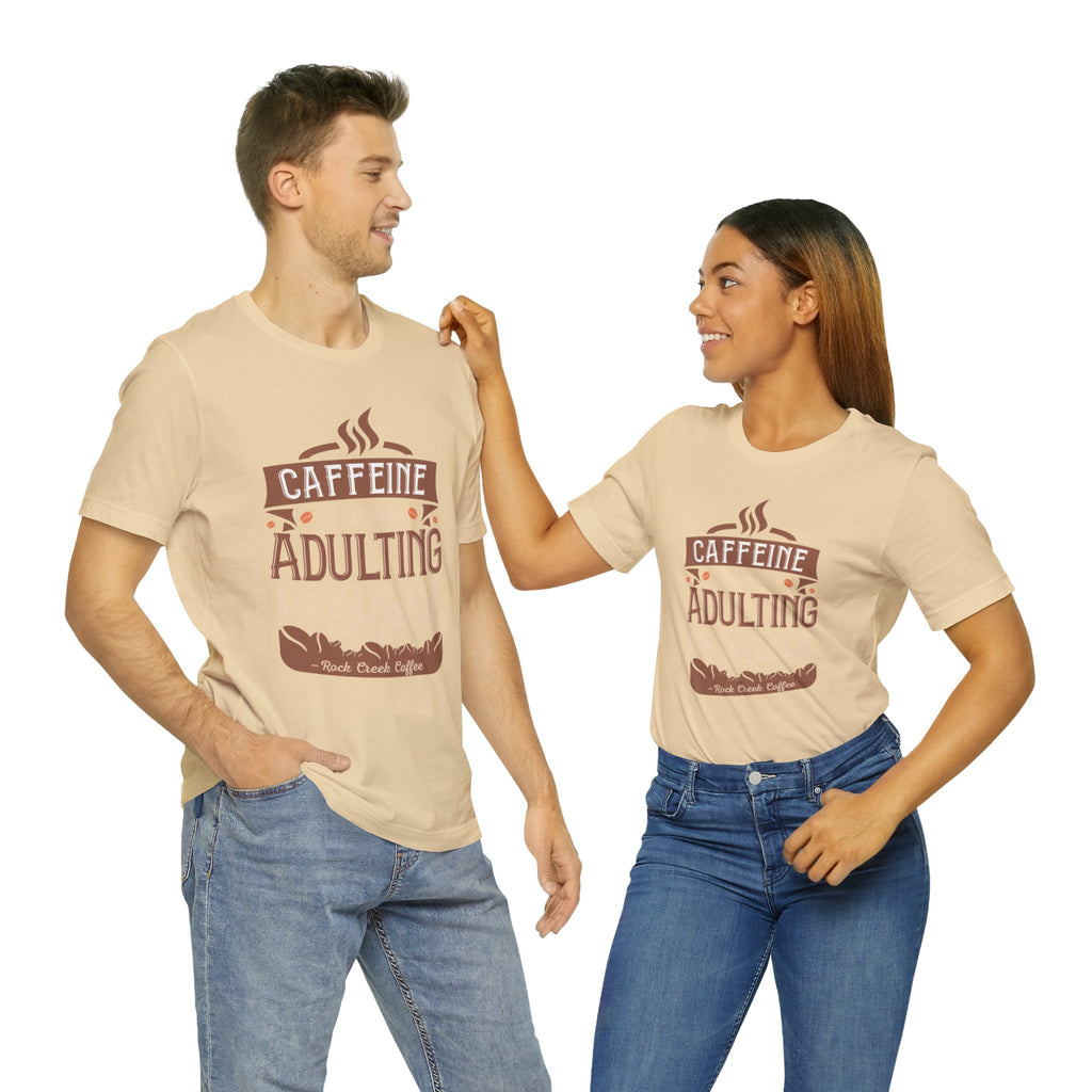 Adult man and woman modeling matching soft cream short-sleeve T-shirts featuring the “Caffeine Because Adulting Is Hard” graphic in a stacked typographic design, posed together against a plain white background.