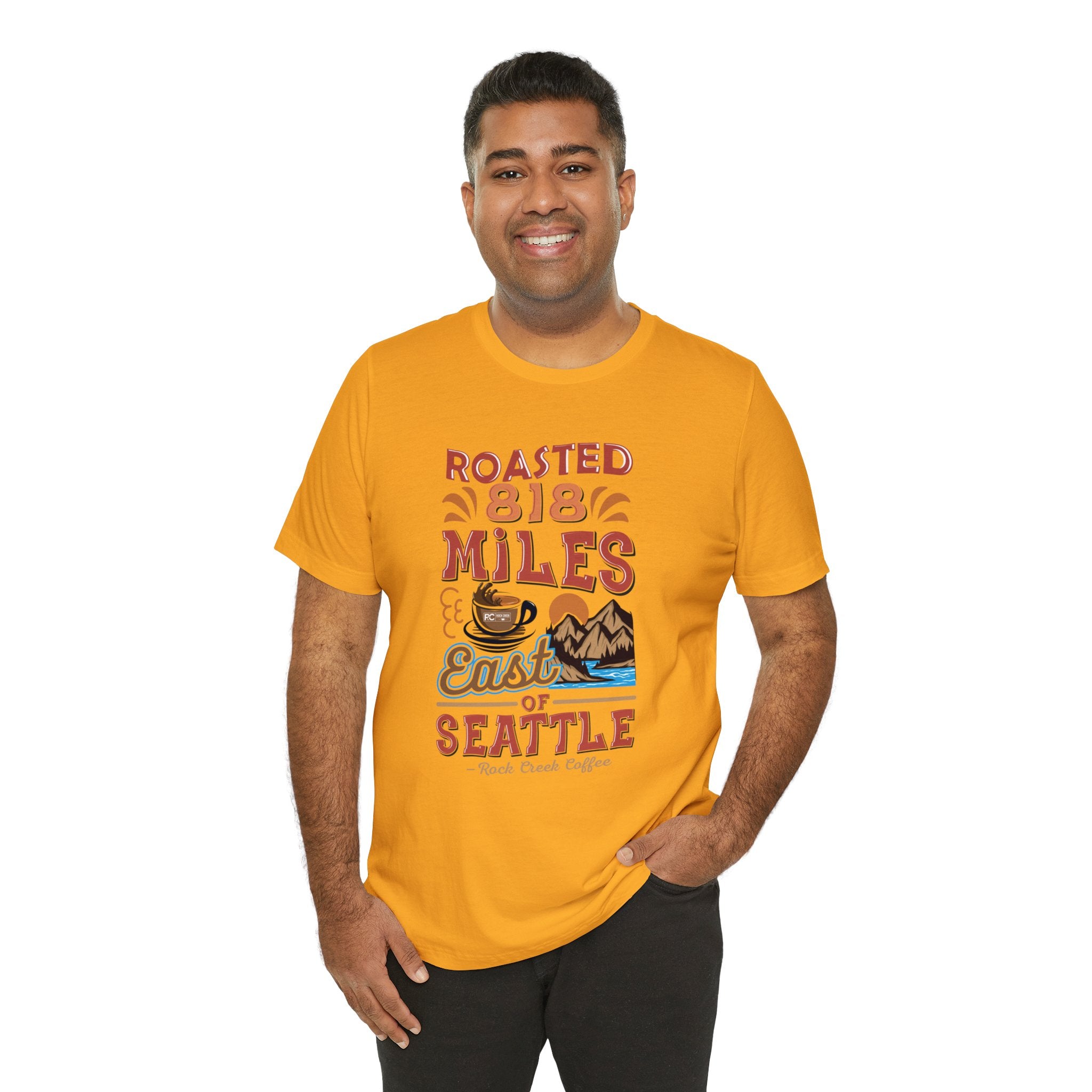 Smiling man modeling a mustard yellow short-sleeve T-shirt printed with a colorful graphic reading “Roasted 818 Miles East of Seattle,” featuring a steaming coffee cup and scenic mountain lake illustration, shown against a plain white background.