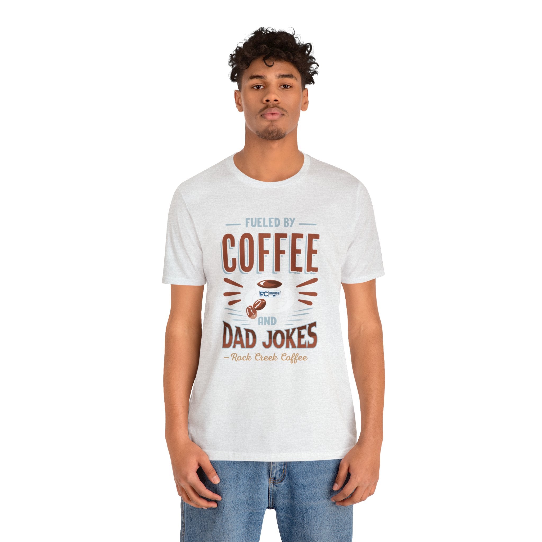 Adult man modeling an ash short-sleeve T-shirt featuring the “Fueled by Coffee and Dad Jokes” graphic with a coffee cup illustration and Rock Creek Coffee text, shown against a plain white background.