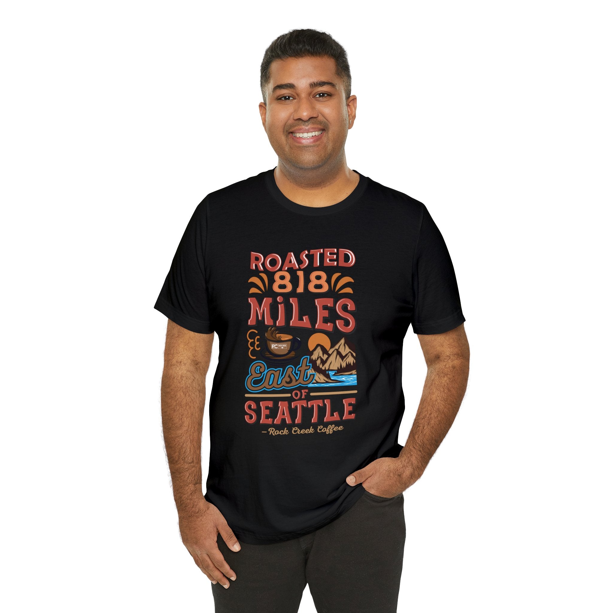 Smiling man modeling a black short-sleeve T-shirt printed with a colorful graphic reading “Roasted 818 Miles East of Seattle,” featuring a steaming coffee cup and a mountain lake illustration, shown against a plain white background.