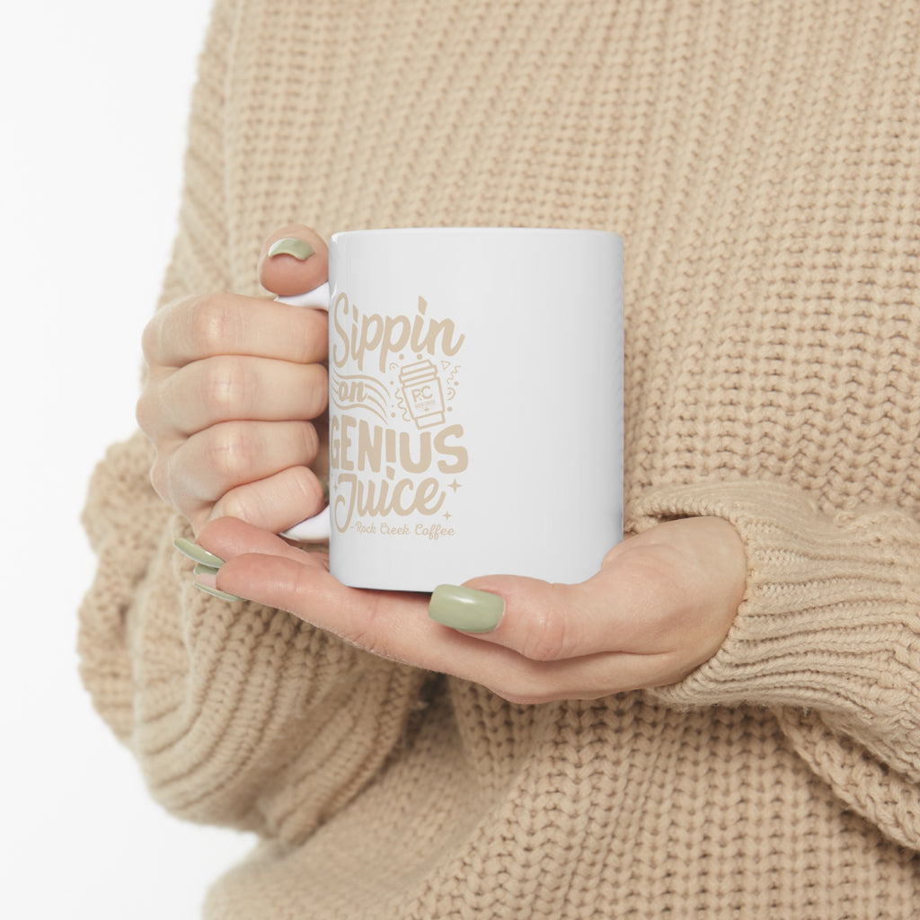 Person wearing a beige knit sweater holding a white Rock Creek Coffee mug with “Sippin on Genius Juice” printed in soft beige lettering and an RC coffee cup graphic.