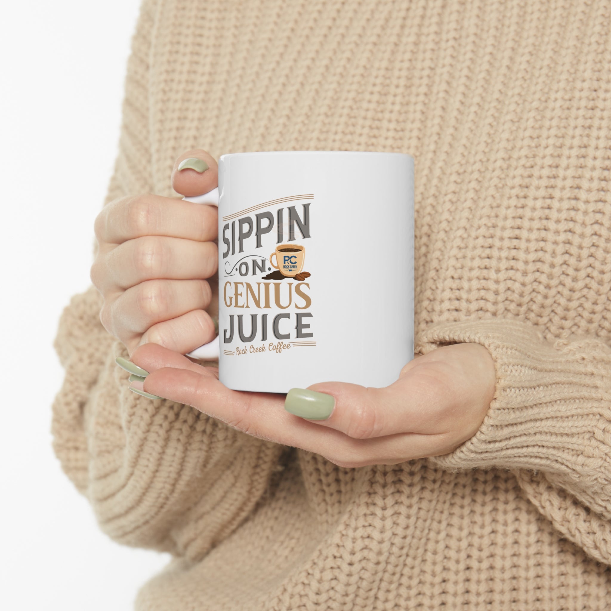 White Rock Creek Coffee mug featuring the phrase “Sippin on Genius Juice” in gray and gold lettering with a small RC coffee cup graphic, held by a person wearing a beige knit sweater with green painted nails against a plain white background.