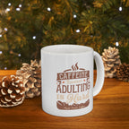 White Rock Creek Coffee mug featuring the phrase “Caffeine Because Adulting Is Hard” in brown and tan lettering, styled on a wooden surface with pinecones and warm holiday lights in the background.