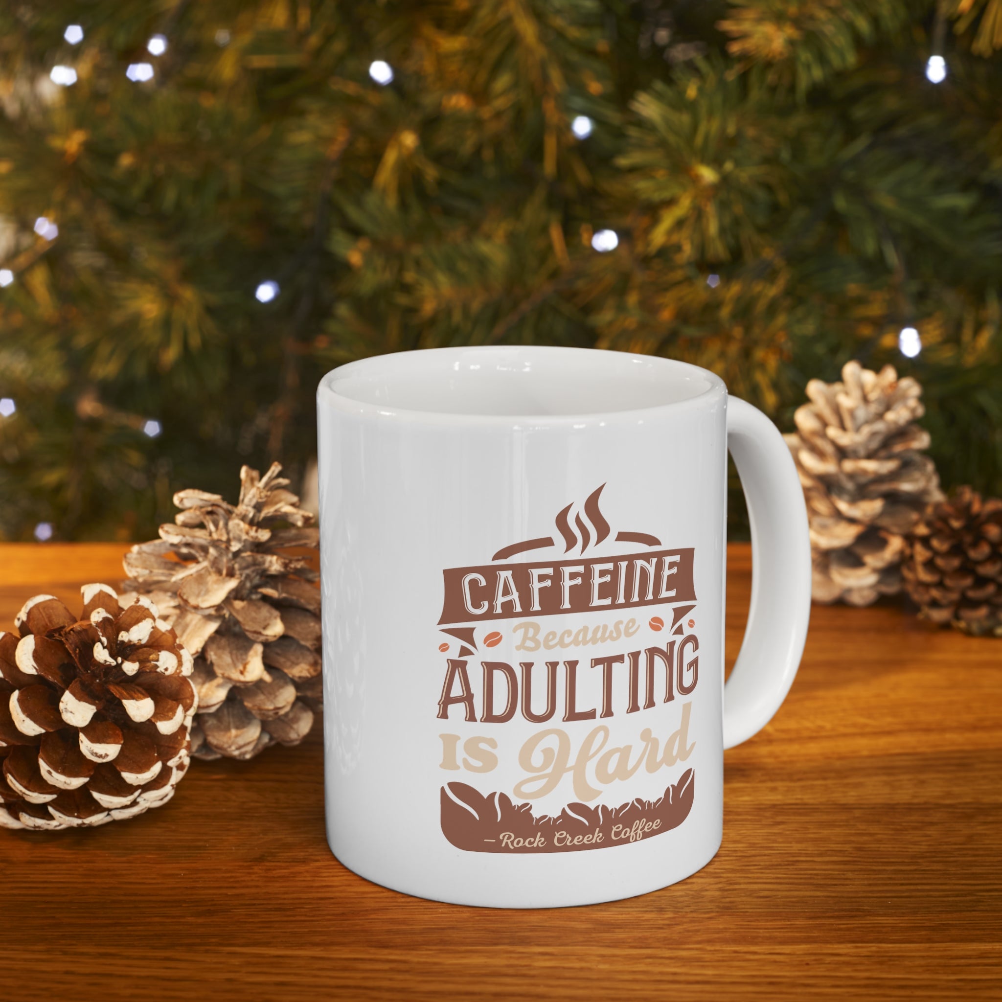 White Rock Creek Coffee mug featuring the phrase “Caffeine Because Adulting Is Hard” in brown and tan lettering, styled on a wooden surface with pinecones and warm holiday lights in the background.