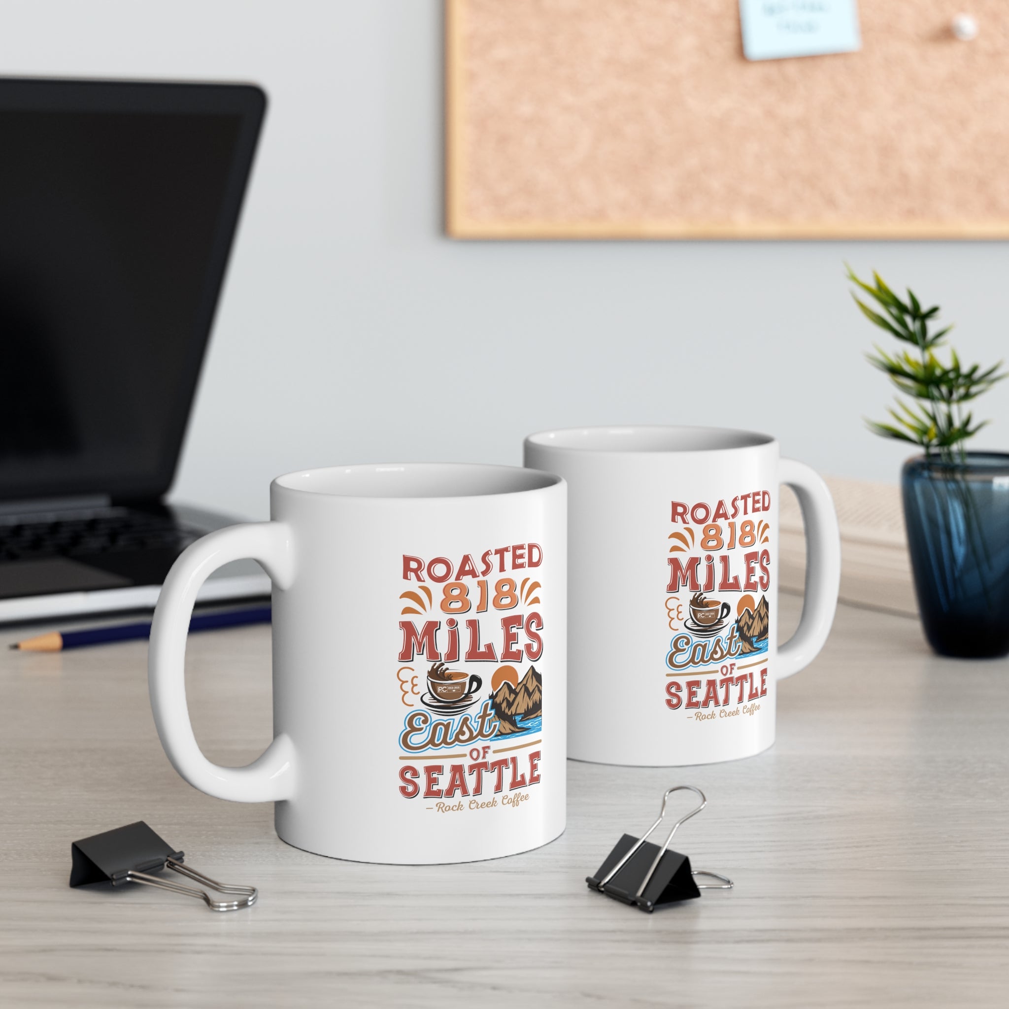 Two white Rock Creek Coffee mugs featuring the colorful “Roasted 818 Miles East of Seattle” design, displayed on a desk with a laptop, binder clips, books, and a small potted plant in the background.