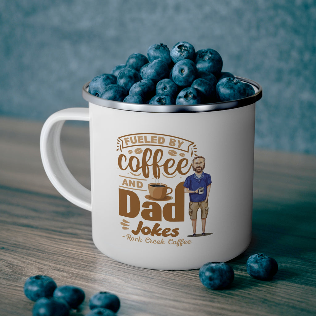 White enamel style coffee mug with silver rim filled with fresh blueberries, featuring the phrase Fueled by Coffee and Dad Jokes Rock Creek Coffee in brown lettering with a coffee cup graphic and illustration of a bearded dad holding a mug, set on a wooden surface with a blue textured background.
