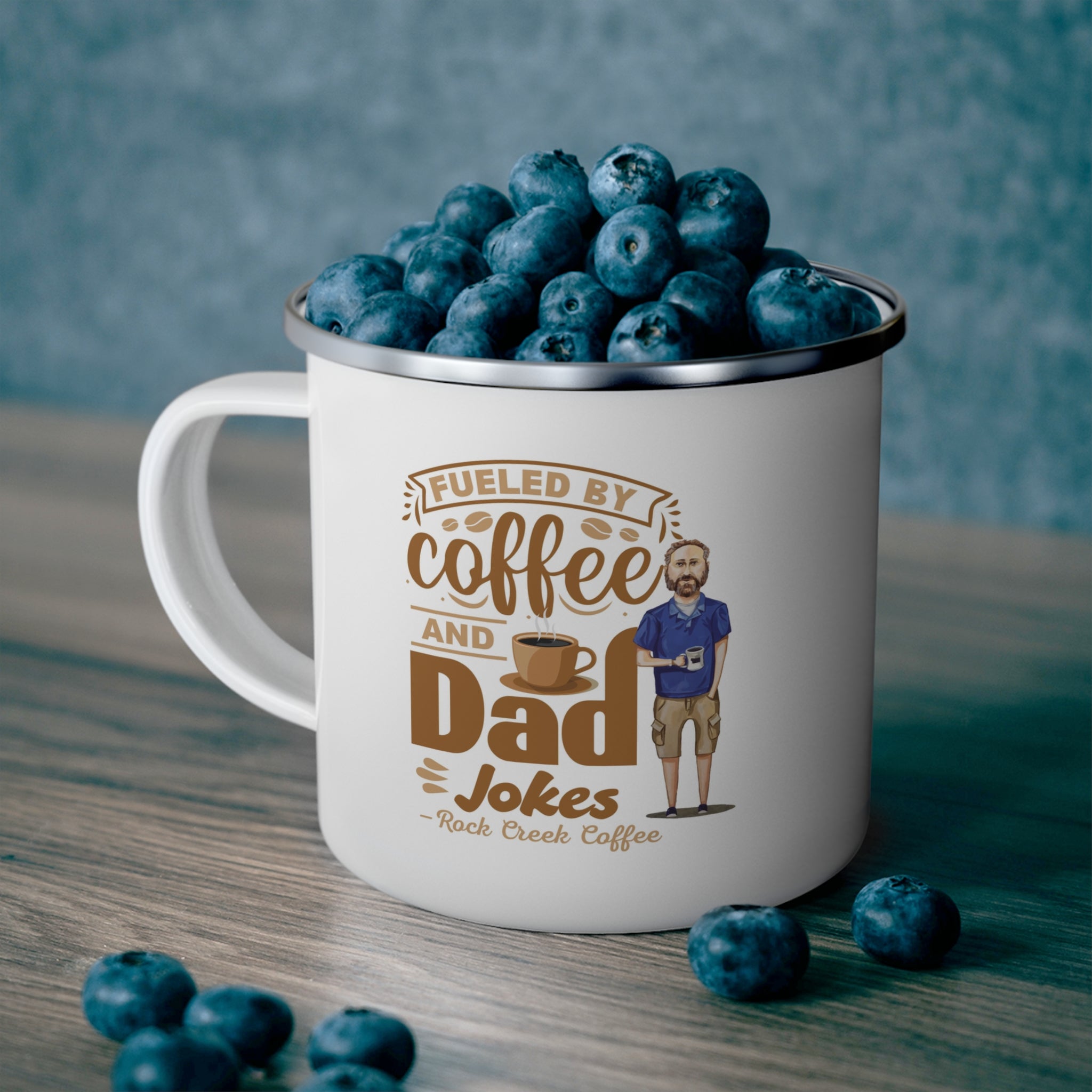 White enamel style coffee mug with silver rim filled with fresh blueberries, featuring the phrase Fueled by Coffee and Dad Jokes Rock Creek Coffee in brown lettering with a coffee cup graphic and illustration of a bearded dad holding a mug, set on a wooden surface with a blue textured background.