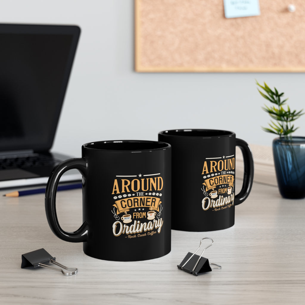 Two black ceramic coffee mugs featuring the Around the Corner from Ordinary Rock Creek Coffee design in gold and white lettering, displayed on a desk with a laptop, binder clips, notebook, and a small potted plant in the background.