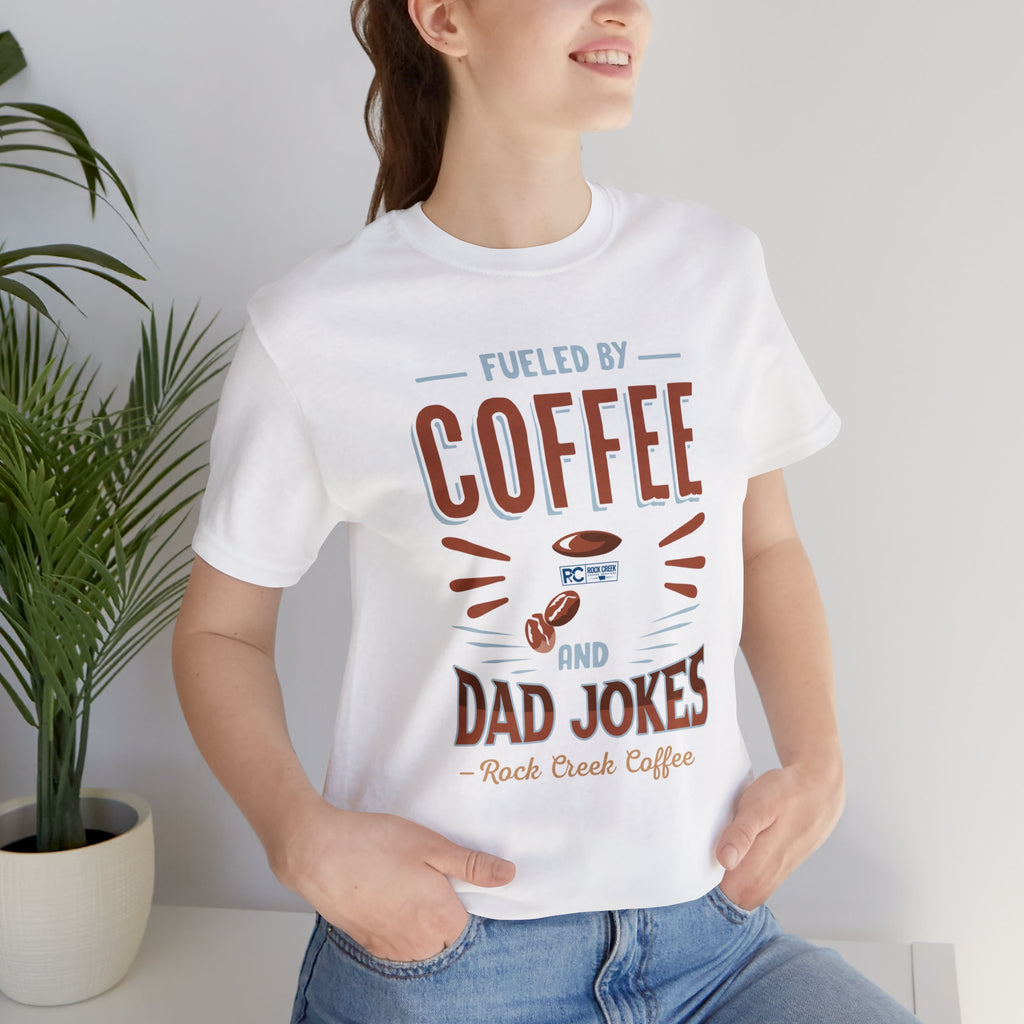 Adult woman modeling a white short-sleeve T-shirt featuring the “Fueled by Coffee and Dad Jokes” graphic with a coffee cup illustration and Rock Creek Coffee text, photographed indoors beside a potted plant against a light neutral background.
