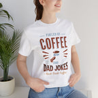 Adult woman modeling a white short-sleeve T-shirt featuring the “Fueled by Coffee and Dad Jokes” graphic with a coffee cup illustration and Rock Creek Coffee text, photographed indoors beside a potted plant against a light neutral background.