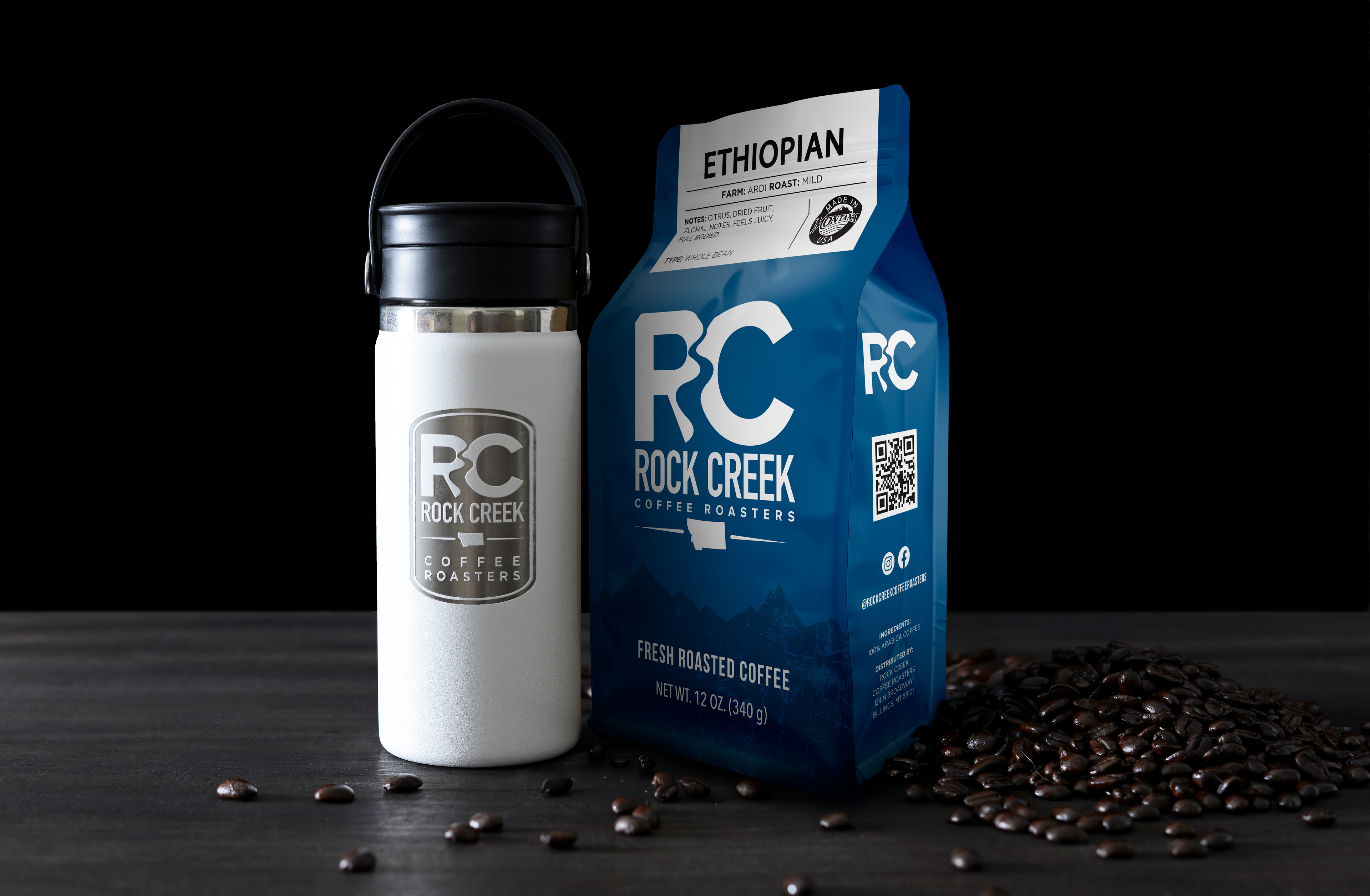 Rock Creek Coffee Roasters Ethiopian Ardi mild roast whole bean coffee bag displayed beside a white insulated travel tumbler with RC logo, with roasted coffee beans scattered on a dark tabletop against a black background.