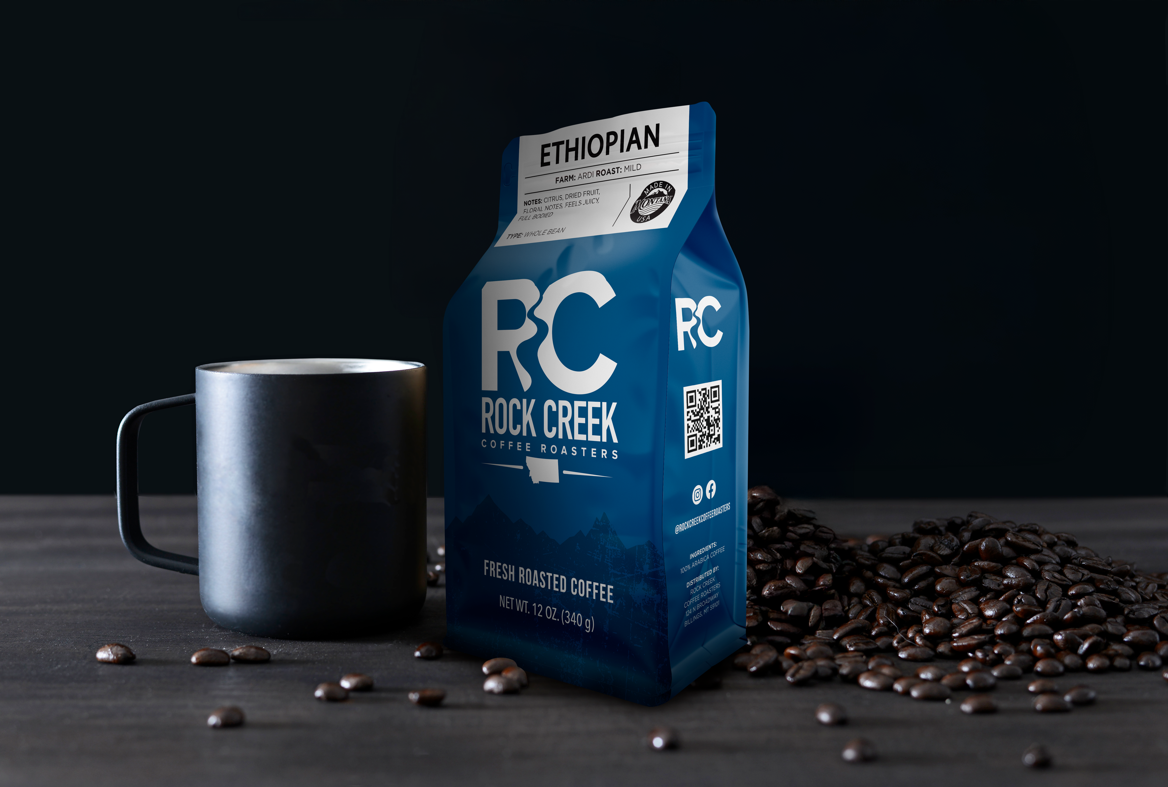 Rock Creek Coffee Roasters Ethiopian Ardi mild roast whole bean coffee bag displayed next to a black ceramic mug, with roasted coffee beans scattered across a dark tabletop against a black background.