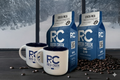 Two Rock Creek Coffee Roasters Costa Rica whole bean coffee bags displayed beside two white RC logo mugs, with roasted coffee beans scattered on a dark tabletop and a snowy mountain landscape visible through large windows in the background.