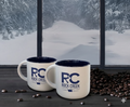 Two white Rock Creek Coffee Roasters RC logo mugs on a dark tabletop with roasted coffee beans scattered nearby, set in front of large windows overlooking a snowy forest landscape.
