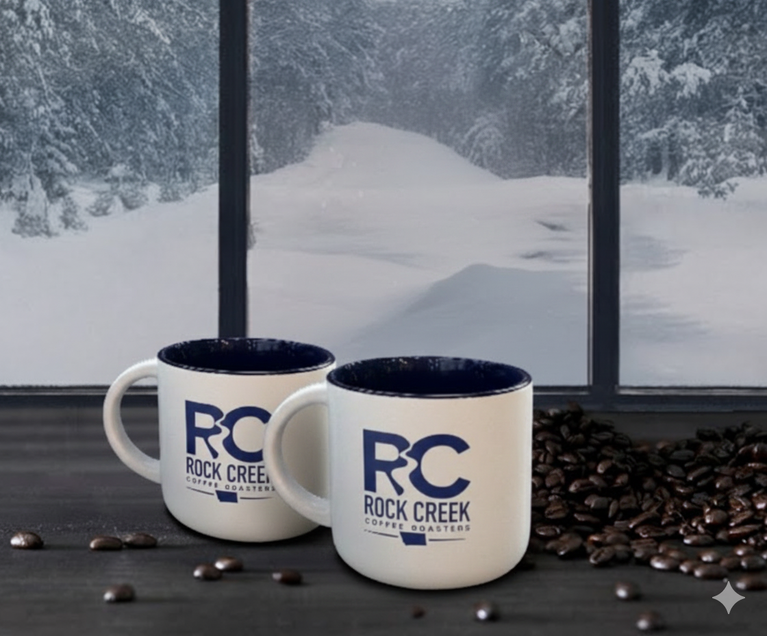 Two white Rock Creek Coffee Roasters RC logo mugs on a dark tabletop with roasted coffee beans scattered nearby, set in front of large windows overlooking a snowy forest landscape.