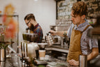Barista in a coffee shop pouring hot water from a stainless steel kettle into a pour-over dripper on a scale, with another barista working in the background and coffee grinders on the counter.