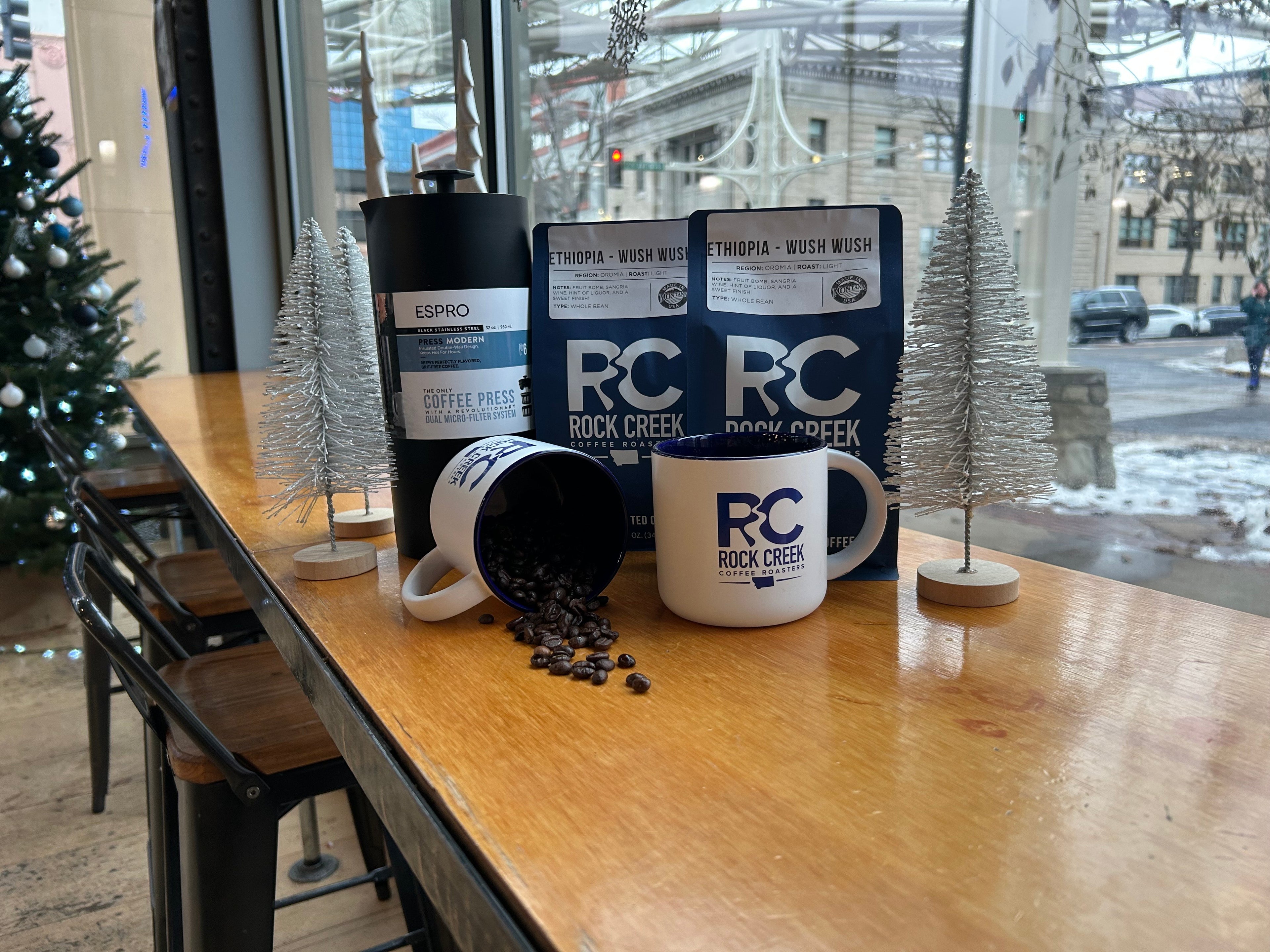 Rock Creek Coffee Roasters display featuring two Ethiopia Wush Wush whole bean coffee bags, an Espro coffee press, and two white RC logo mugs on a wooden table by a window, with one mug tipped over spilling roasted coffee beans, surrounded by small decorative white trees and a snowy street scene outside.