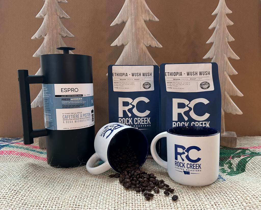 Rock Creek Coffee Roasters display featuring two Ethiopia Wush Wush whole bean coffee bags, an Espro coffee press, and two white RC logo mugs, with one mug tipped over spilling roasted coffee beans, arranged on burlap fabric with wooden tree decorations in the background.