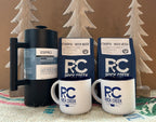 Rock Creek Coffee Roasters display featuring two Ethiopia Wush Wush whole bean coffee bags, an Espro coffee press, and two white RC logo mugs arranged on burlap fabric, with wooden tree decorations in the background.