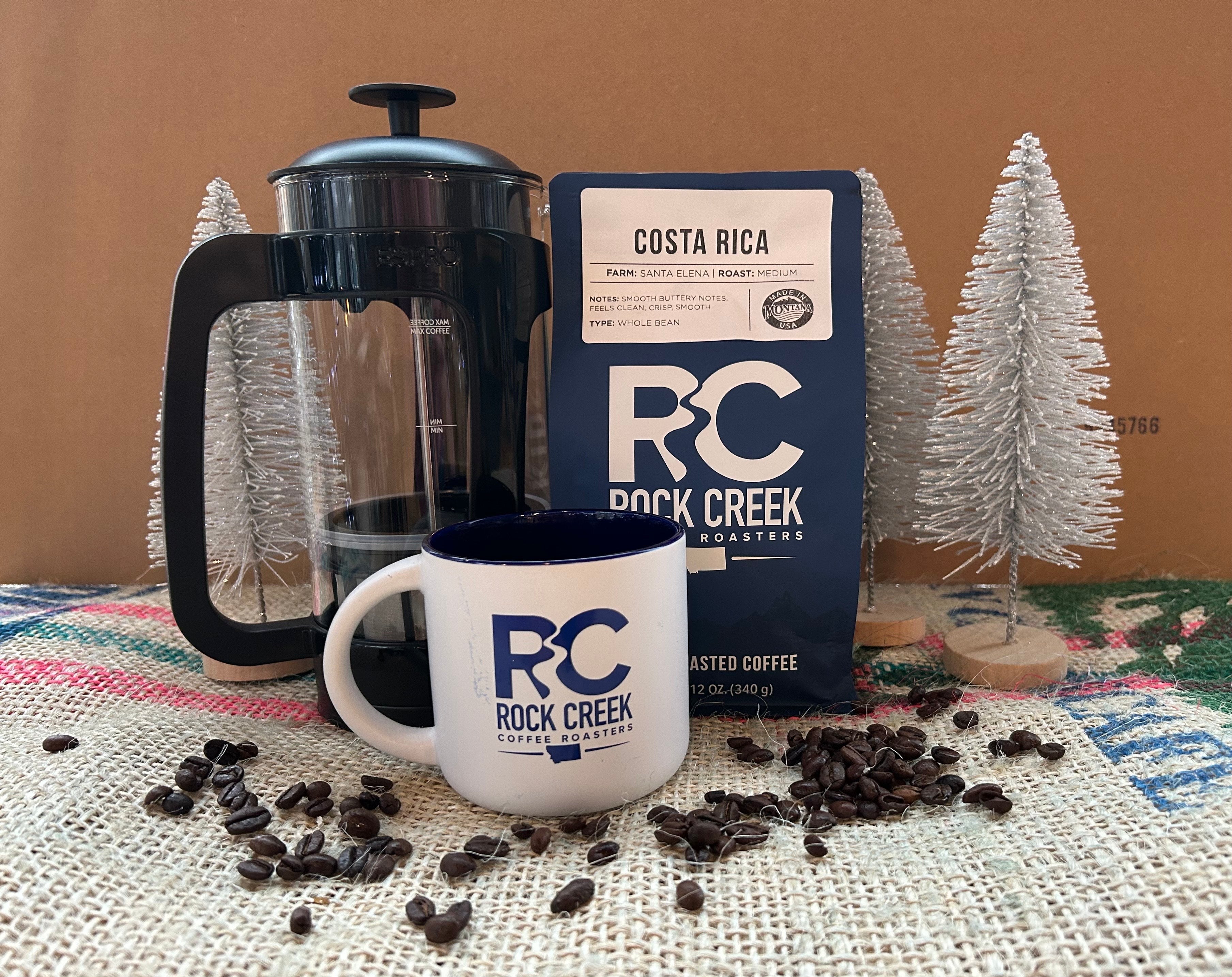 Rock Creek Coffee Roasters Costa Rica Santa Elena medium roast whole bean coffee bag displayed with a French press and a white RC logo mug, surrounded by scattered roasted coffee beans on burlap fabric, with small decorative white trees in the background.