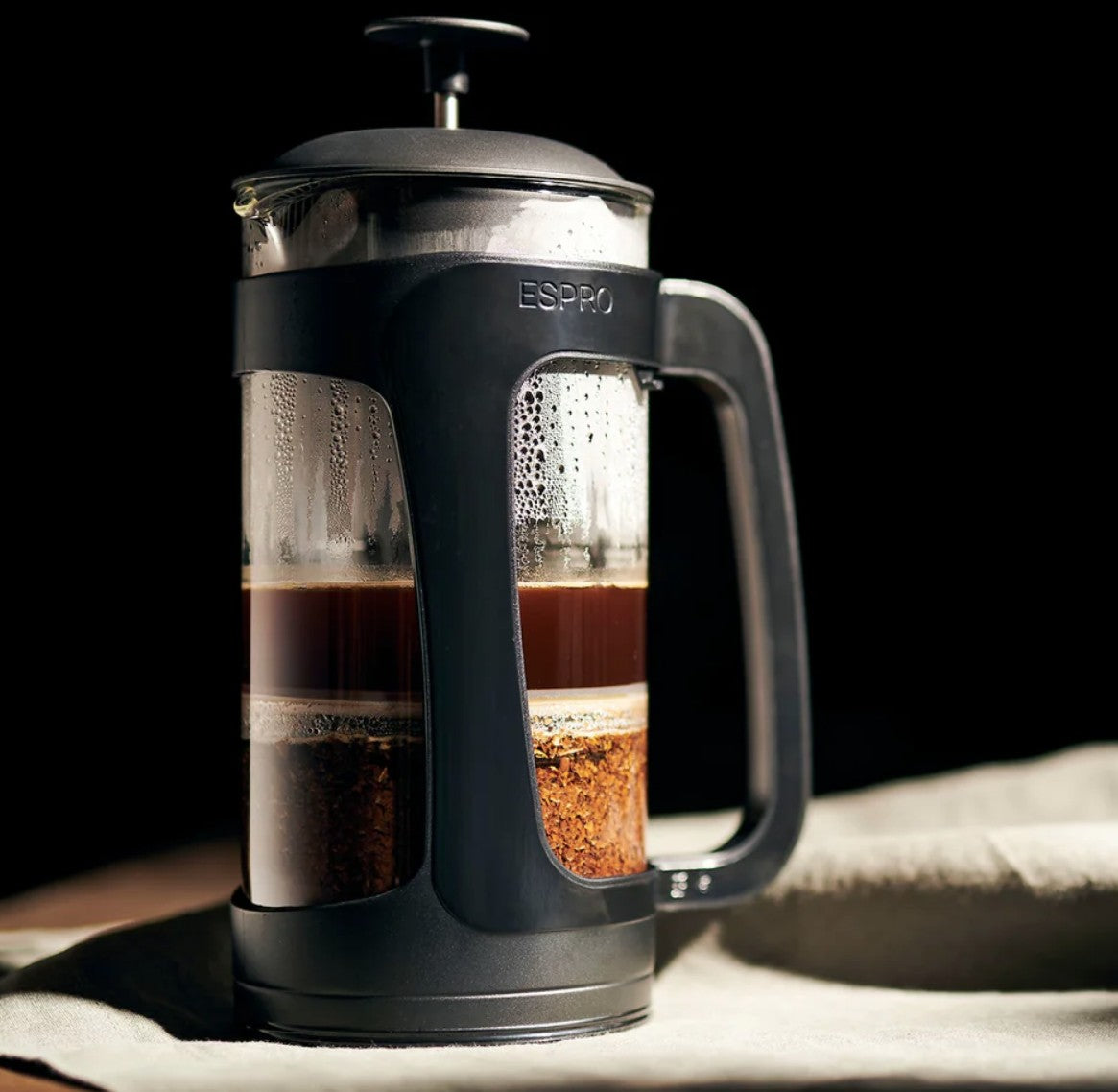 ESPRO French press brewing coffee with visible layers of grounds and dark coffee inside a glass carafe, black frame and handle, condensation on the glass, set against a dark background.