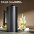 Matte black stainless steel French press coffee maker with handle and plunger on a countertop, with a glass jar of iced tea and rosemary in the background and text reading “Durable Stainless Steel” and “Brews in 4 Minutes.”