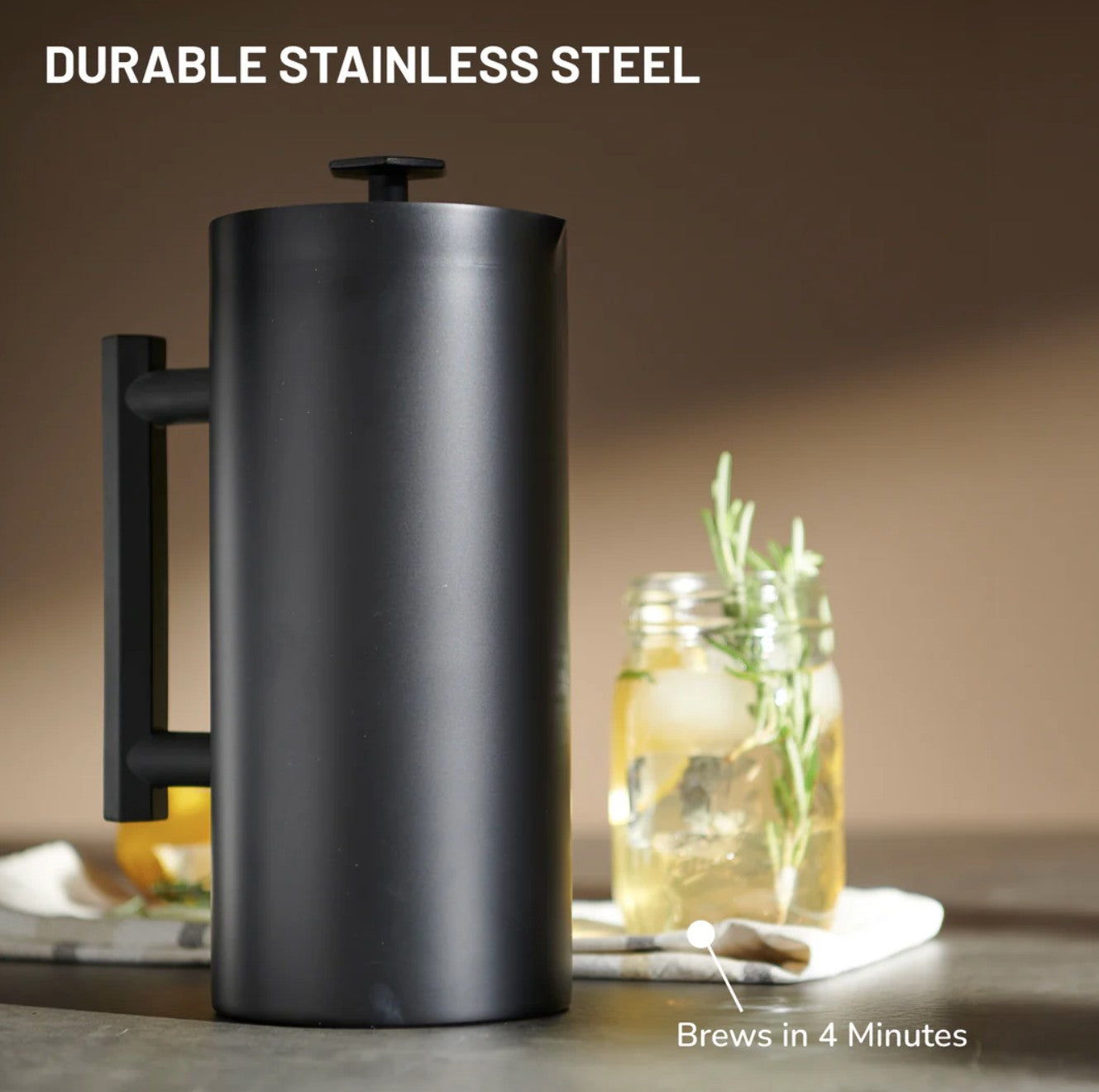 Matte black stainless steel French press coffee maker with handle and plunger on a countertop, with a glass jar of iced tea and rosemary in the background and text reading “Durable Stainless Steel” and “Brews in 4 Minutes.”