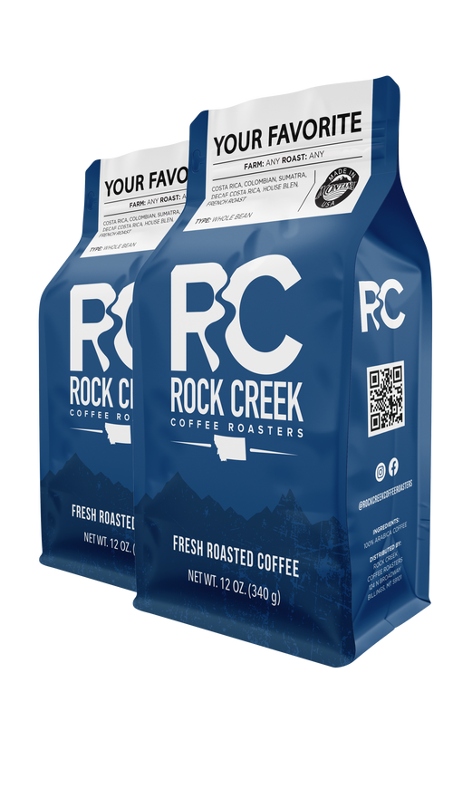 Two blue 12-oz bags of Rock Creek Coffee Roasters whole bean coffee labeled “Your Favorite,” featuring the RCC logo and Montana-inspired mountain design
