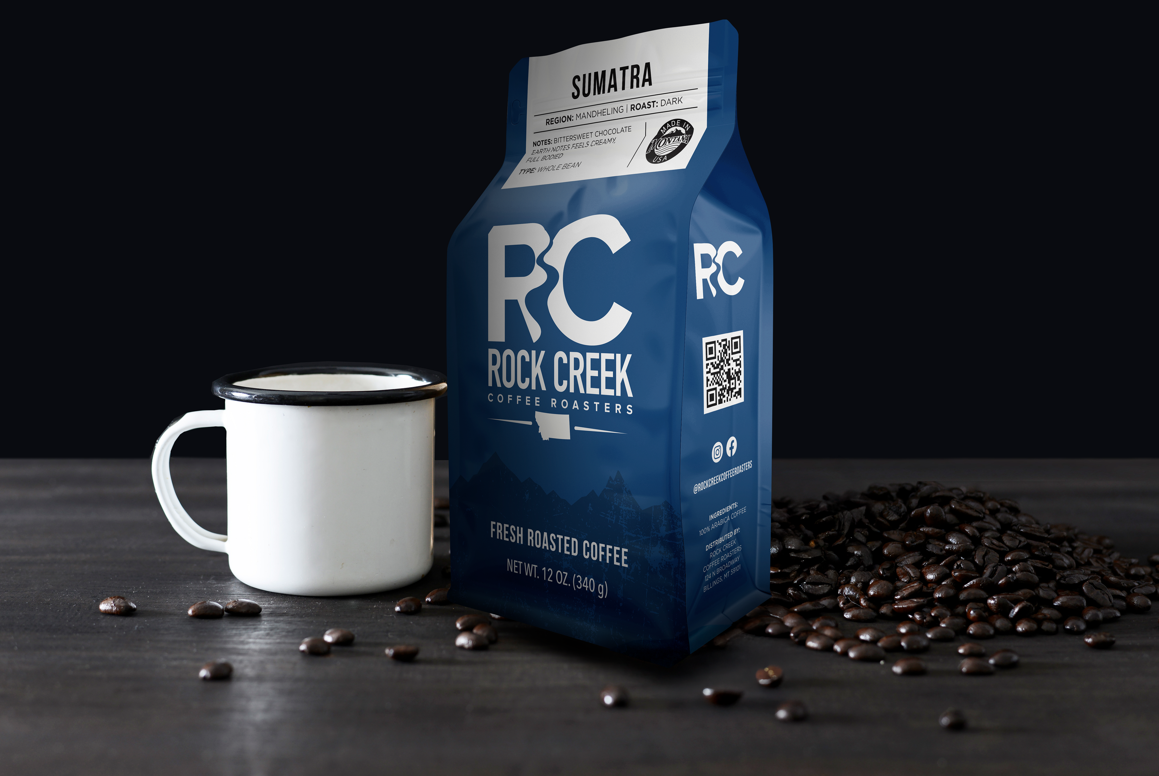 Rock Creek Coffee Roasters Sumatra Mandheling dark roast whole bean coffee bag displayed beside a white enamel mug, with roasted coffee beans scattered on a dark tabletop against a black background.