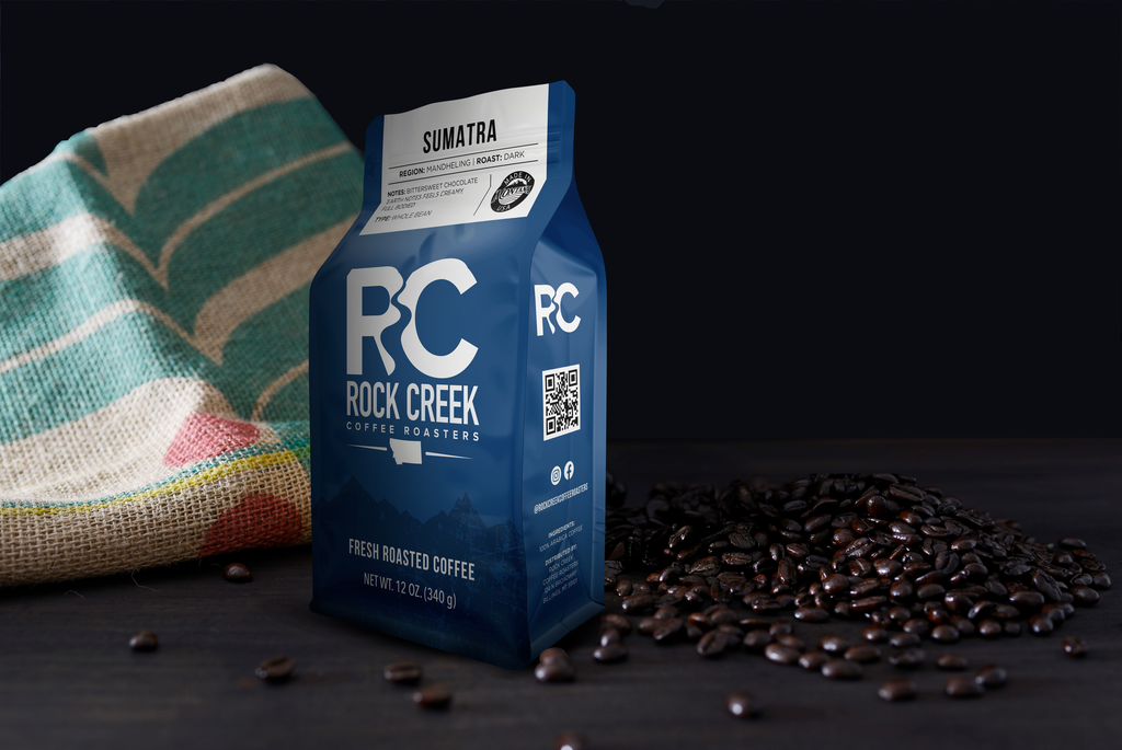Rock Creek Coffee Roasters Sumatra Mandheling dark roast whole bean coffee bag displayed on a dark tabletop with a pile of roasted coffee beans and a burlap coffee sack in the background against a black backdrop.