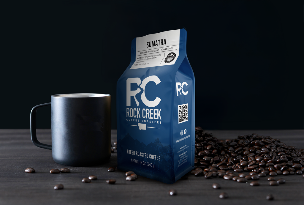 Rock Creek Coffee Roasters Sumatra Mandheling dark roast whole bean coffee bag displayed beside a matte black mug, with roasted coffee beans scattered on a dark tabletop against a black background.