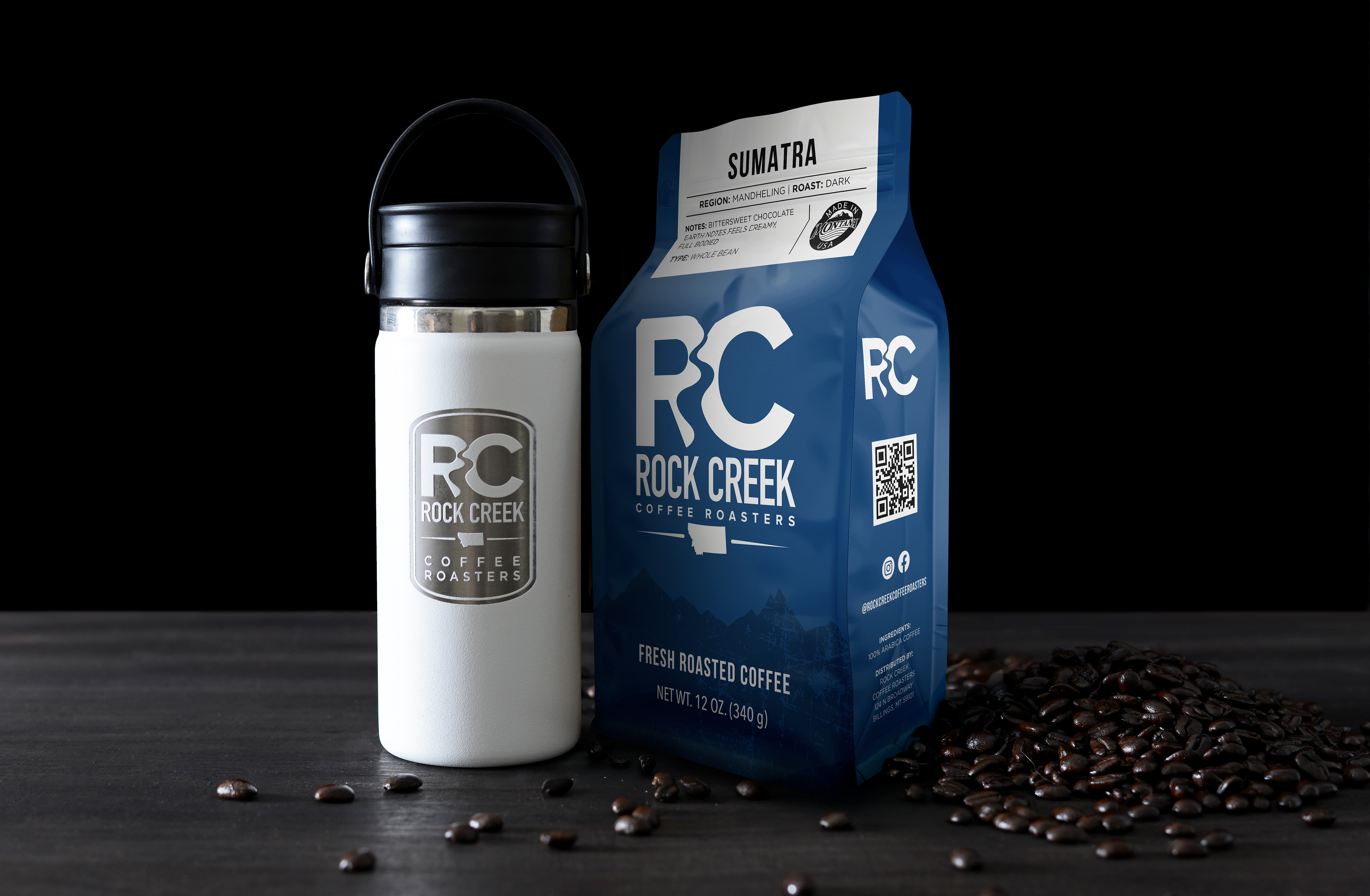 Rock Creek Coffee Roasters Sumatra Mandheling dark roast whole bean coffee bag displayed beside a white insulated travel tumbler with RC logo, with roasted coffee beans scattered on a dark tabletop against a black background.