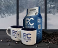 Rock Creek Coffee Roasters Costa Rica whole bean coffee bag displayed with two white RC logo mugs and scattered roasted coffee beans on a dark table, set in front of large windows overlooking a snowy forest landscape.