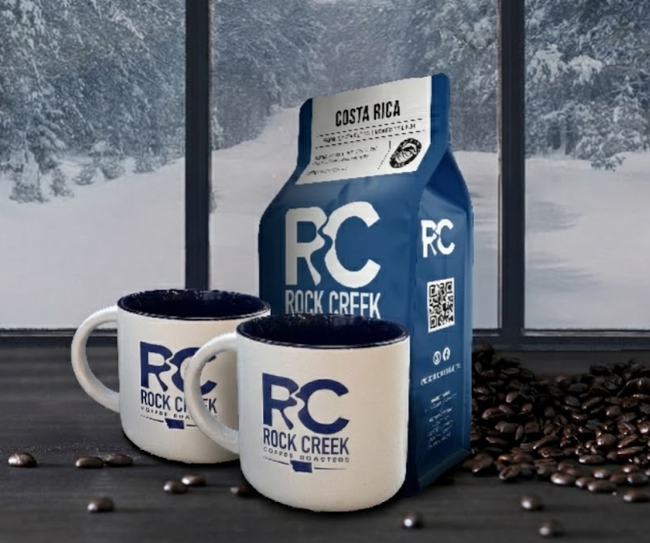 Rock Creek Coffee Roasters Costa Rica whole bean coffee bag displayed with two white RC logo mugs and scattered roasted coffee beans on a dark table, set in front of large windows overlooking a snowy forest landscape.