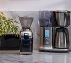 Baratza Virtuoso Plus coffee grinder next to a Breville Precision Brewer coffee maker on a marble countertop, with stainless steel thermal carafe and a leafy houseplant in the background.