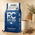 Rock Creek Coffee Roasters Colombian coffee bag beside a steaming cup of coffee and scattered roasted beans on a warm neutral countertop.