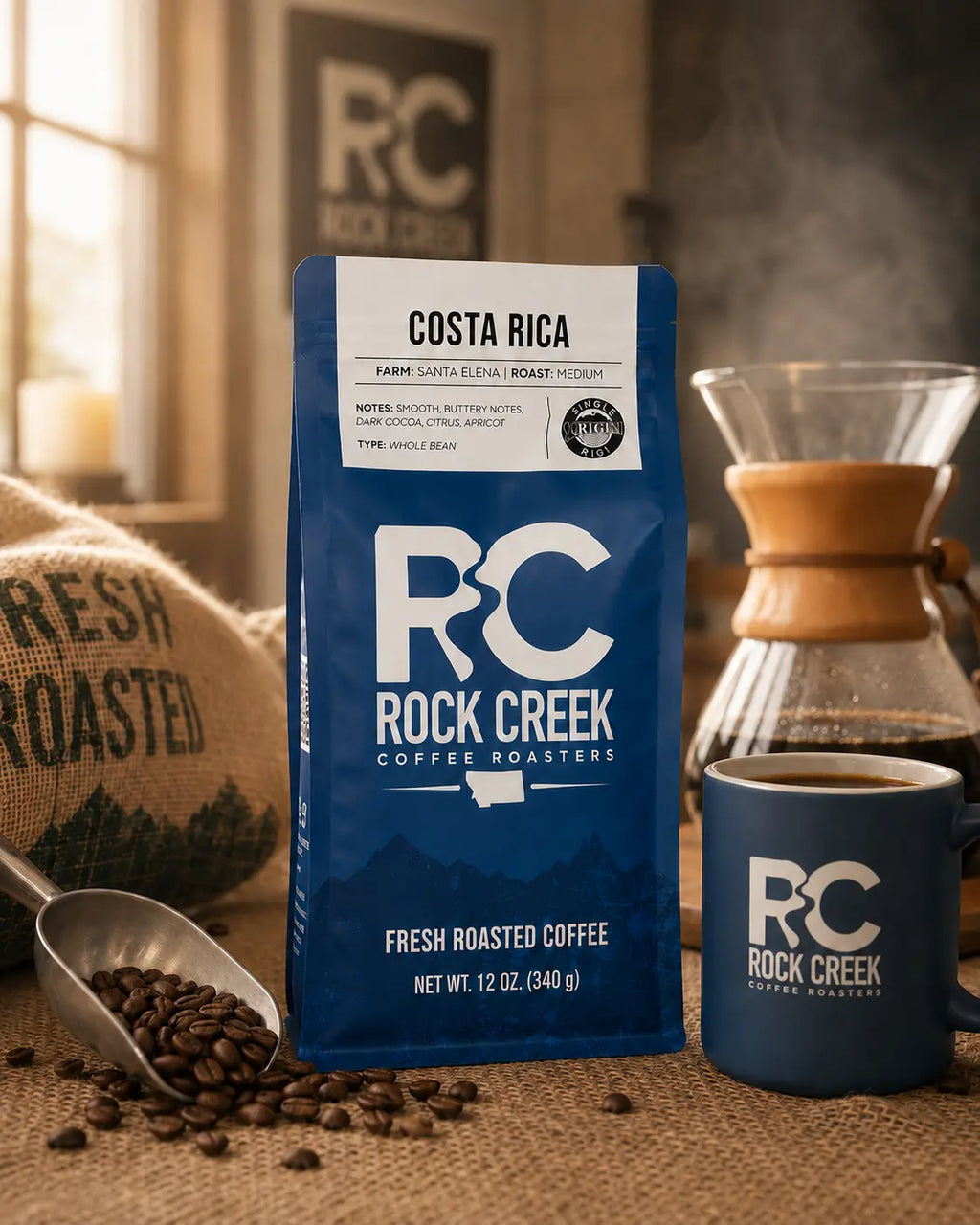Rock Creek Coffee Roasters Costa Rica whole bean coffee bag in a warm roastery-style setting with coffee beans, a scoop, a branded mug, and brewing equipment.