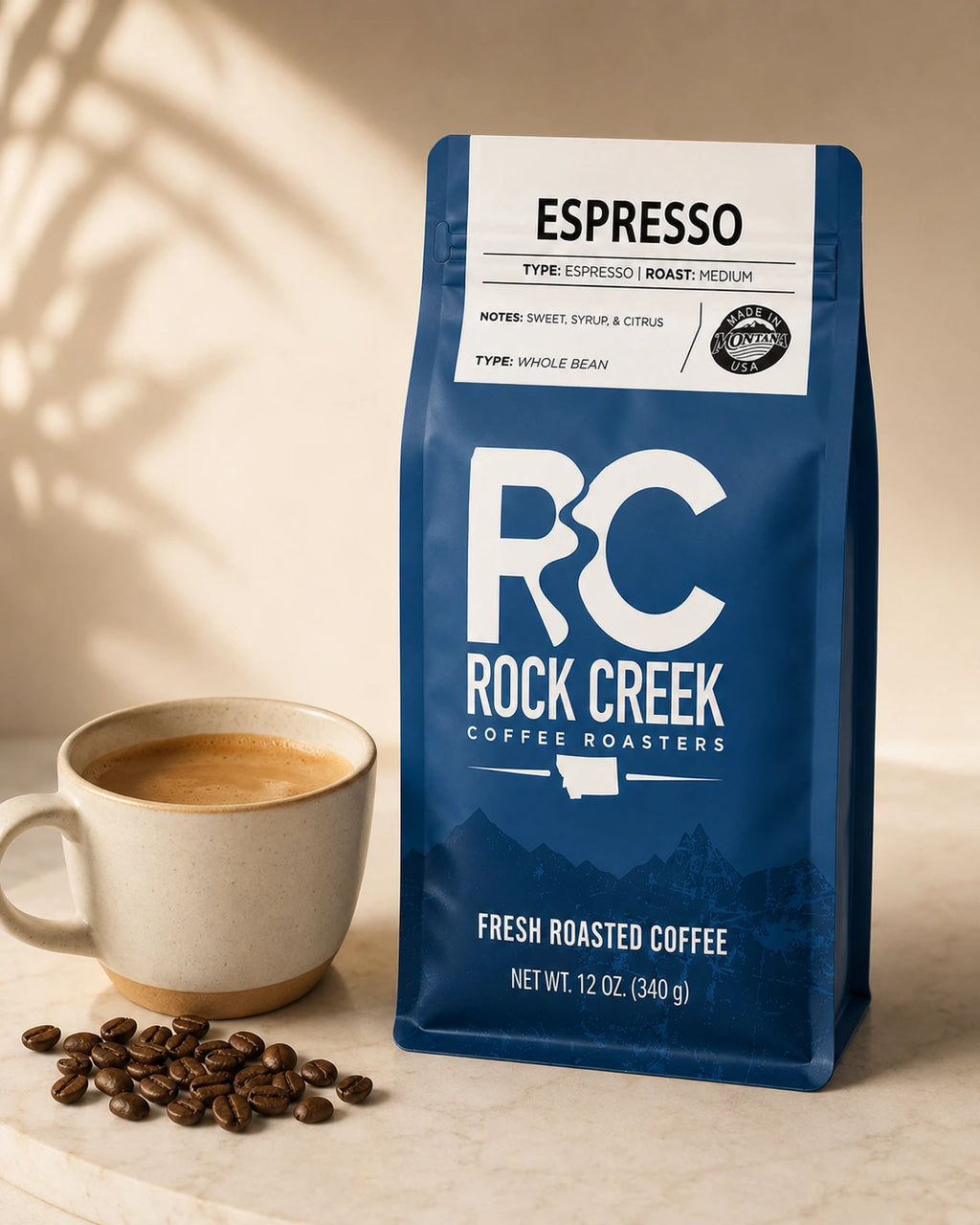 Rock Creek Coffee Roasters Espresso coffee bag beside a cup of brewed coffee and roasted coffee beans on a warm neutral tabletop.