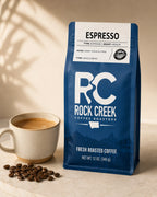 Rock Creek Coffee Roasters Espresso coffee bag beside a cup of brewed coffee and roasted coffee beans on a warm neutral tabletop.