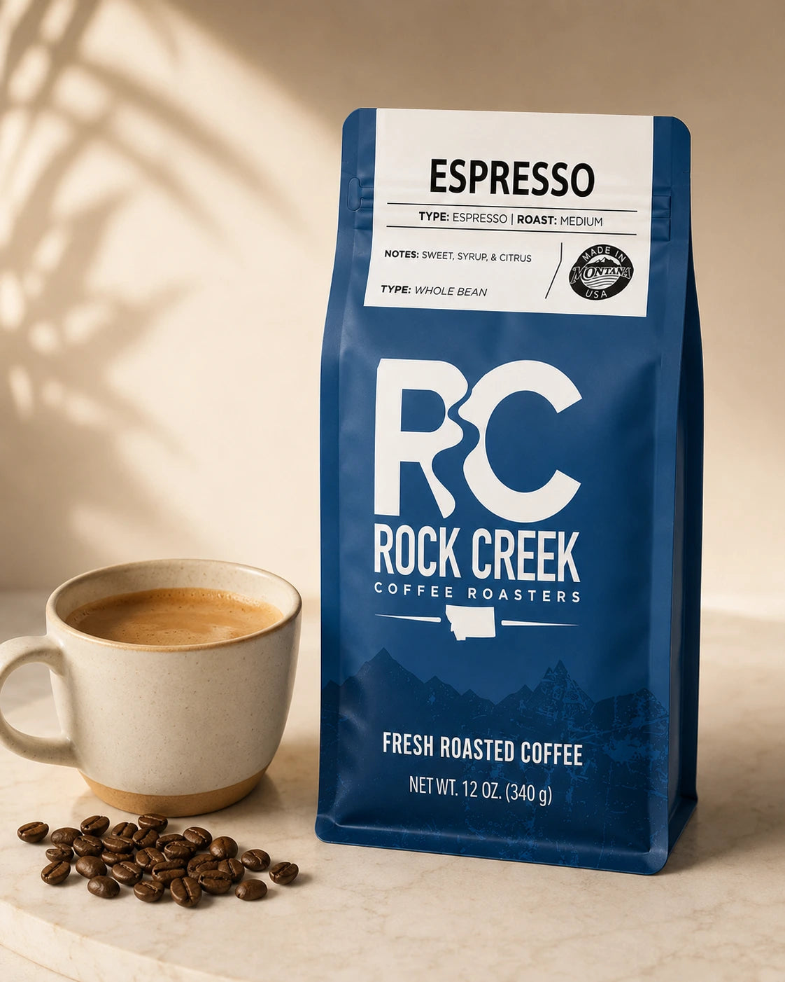 Rock Creek Coffee Roasters Espresso coffee bag beside a cup of brewed coffee and roasted coffee beans on a warm neutral tabletop.