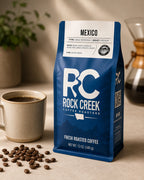 Rock Creek Coffee Roasters Mexico coffee bag displayed on a warm tabletop beside a brewed coffee cup, scattered coffee beans, and a blurred pour-over brewer.