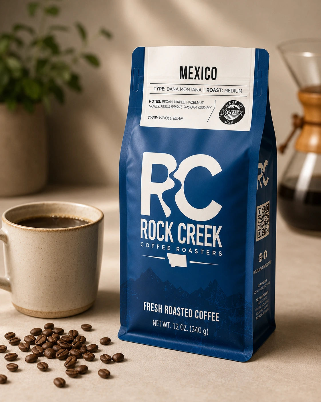 Rock Creek Coffee Roasters Mexico coffee bag displayed on a warm tabletop beside a brewed coffee cup, scattered coffee beans, and a blurred pour-over brewer.