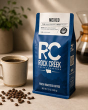 Load image into Gallery viewer, Rock Creek Coffee Roasters Mexico coffee bag displayed on a warm tabletop beside a brewed coffee cup, scattered coffee beans, and a blurred pour-over brewer.