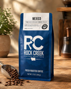 Rock Creek Coffee Roasters Mexico coffee bag displayed in a warm kitchen setting with coffee beans, a scoop, and soft natural lighting.