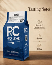 Load image into Gallery viewer, Rock Creek Coffee Roasters Brazil coffee bag displayed on a warm wooden surface with a Tasting Notes graphic highlighting roasted ground nuts, sweet, and toffee flavor notes.